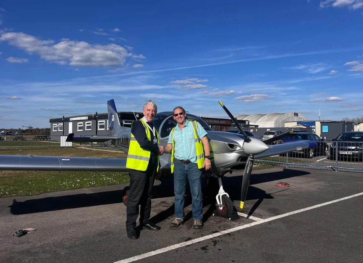 Two men shaking hands in front of a small silver airplane, both wearing high-visibility vests and smiling, in an outdoor parking lot on a sunny day with blue sky and clouds.