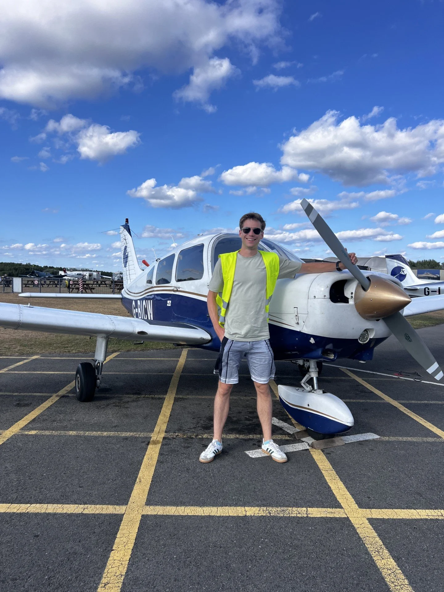 A man wearing sunglasses, a neon yellow safety vest, a light gray t-shirt, and striped shorts standing in front of a small blue and white airplane, holding the propeller with a smile on his face, at an airport tarmac under a partly cloudy sky.