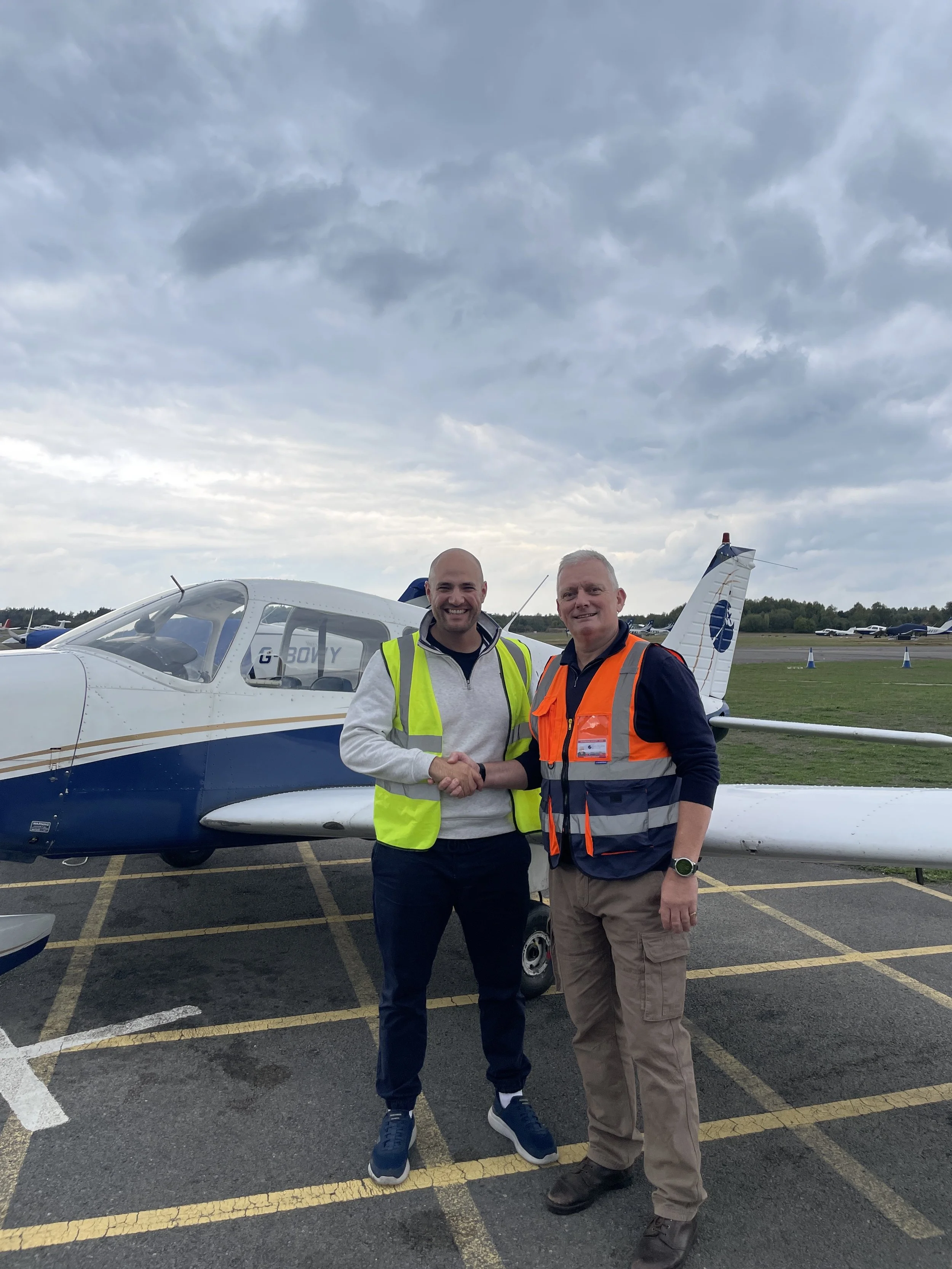 Two men shaking hands in front of a small airplane at an airport, with a cloudy sky overhead.