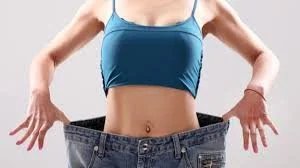 A woman showing off her weight loss while wearing a tank top and pulling open a pair of large, loose-fitting jeans to show a flat stomach.