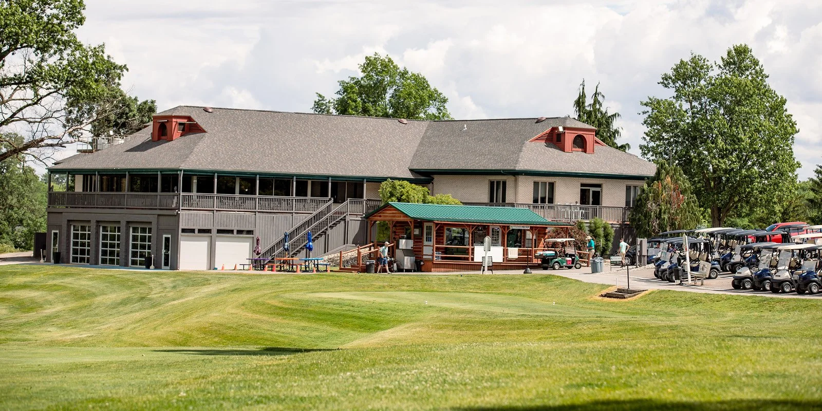 Large building with a gray roof and red accents, surrounded by green trees and a golf course, with golf carts parked nearby.