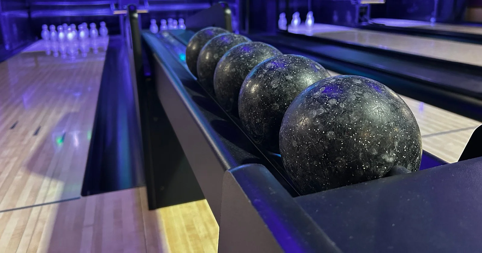 Close-up of five bowling balls on a ball return at a bowling alley, with bowling pins visible in the background.