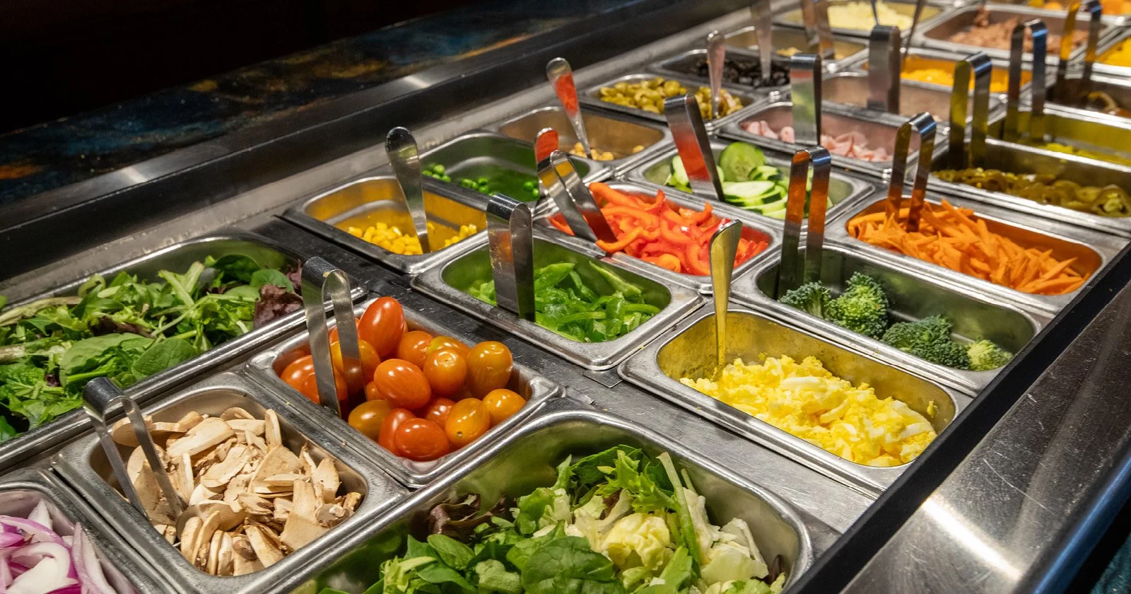 Variety of fresh vegetables in a salad bar, including cherry tomatoes, broccoli, shredded carrots, cucumbers, lettuce, spinach, bell peppers, and shredded cheese, with serving utensils in each container.