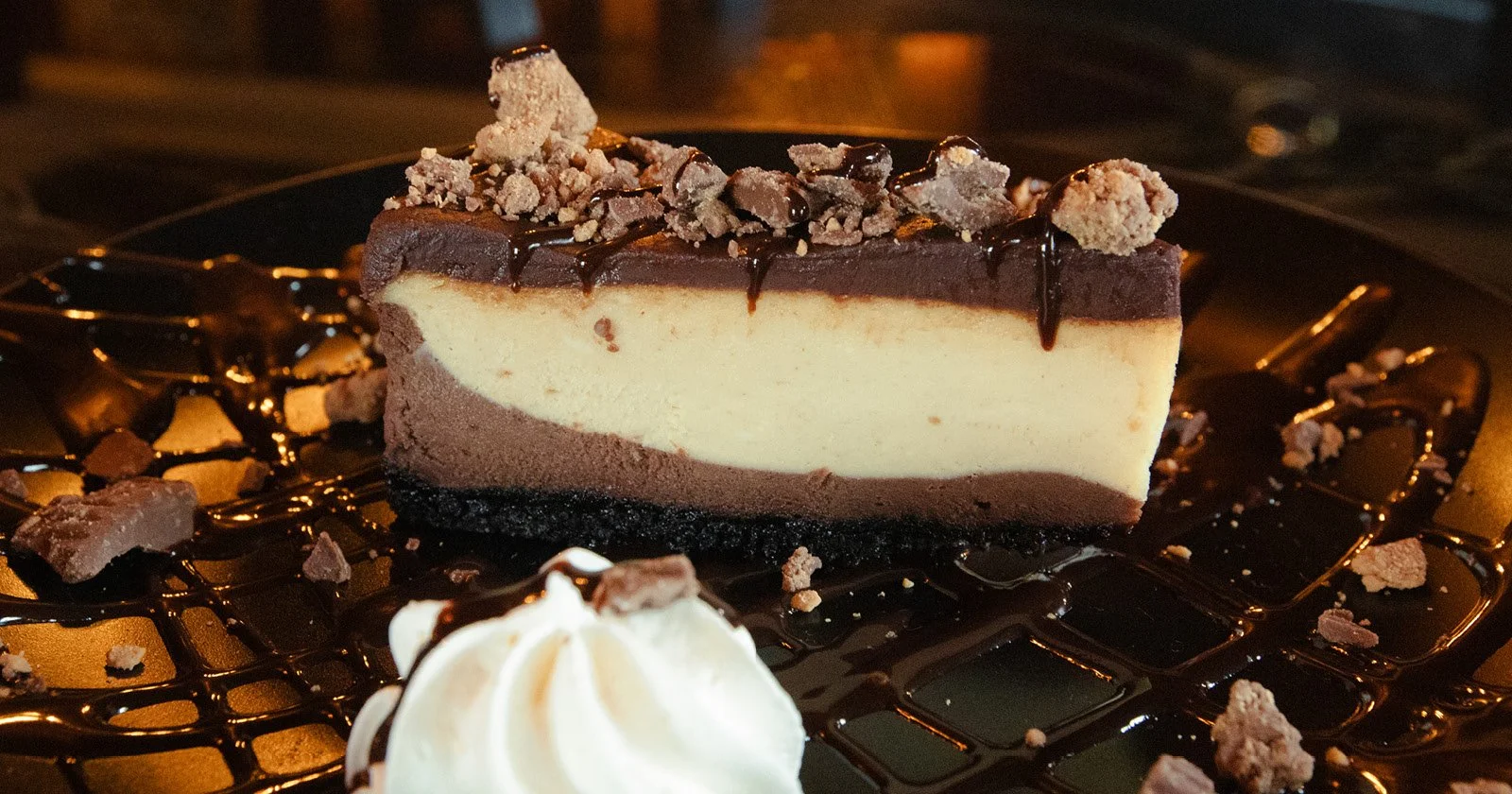 A slice of cheesecake topped with chocolate ganache, crumbled cookies, and chocolate drizzle, served on a black plate with a dollop of whipped cream and surrounded by additional crumbles and chocolate drizzles.