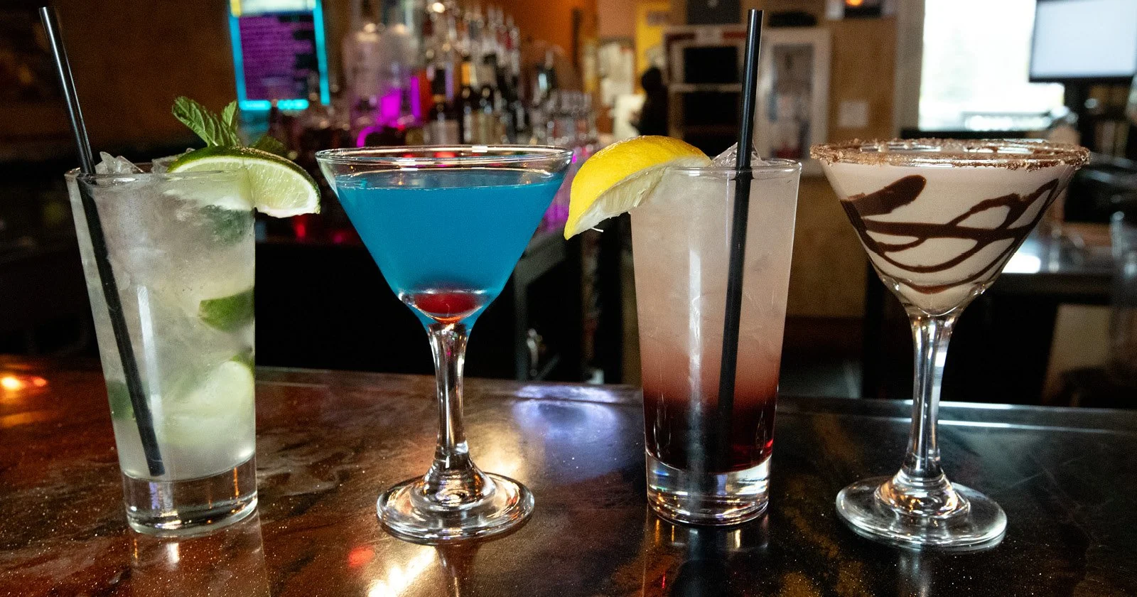 A variety of four colorful alcoholic cocktails on a bar counter, each garnished with lemon or lime slices, with bar equipment and colorful lighting in the background.