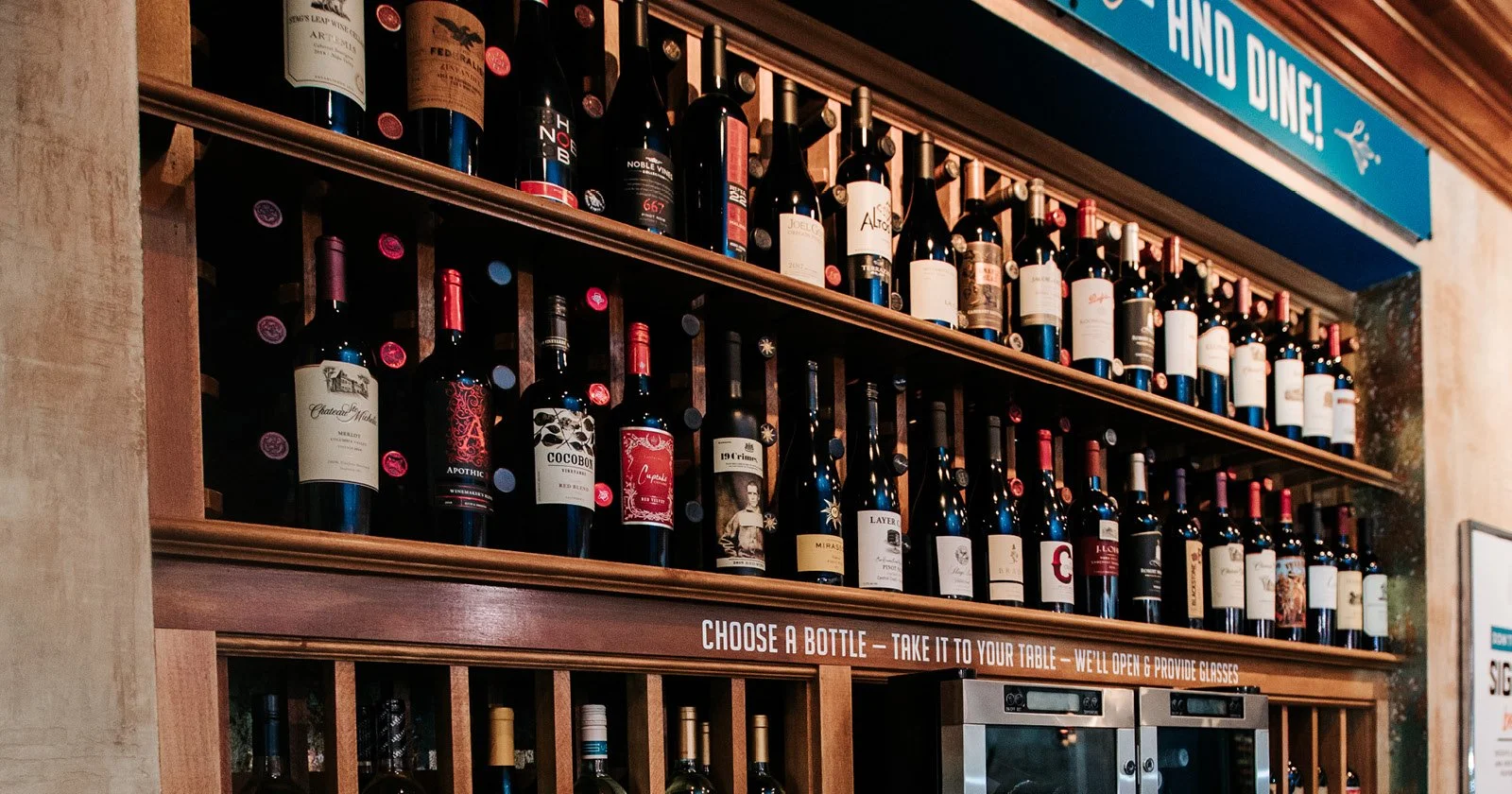 Display of wine bottles on wooden shelves in a restaurant or wine bar, with a blue sign above that says 'HAVE A GLASS AND DINE!' and a counter below with a sign instructing customers to choose a bottle, take it to their table, and that glasses will b