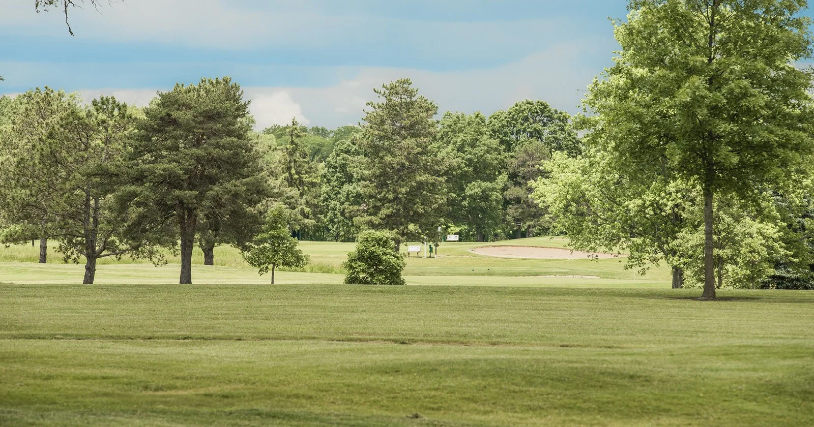 A golf course with well-maintained grass and various trees, under a partly cloudy sky.