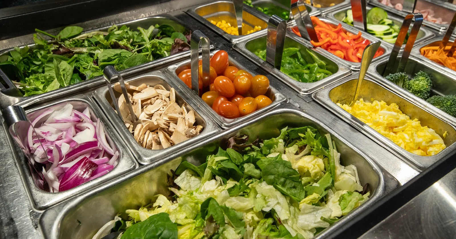 A salad bar with containers of fresh vegetables and toppings, including cherry tomatoes, sliced mushrooms, chopped onions, mixed greens, and sliced bell peppers.