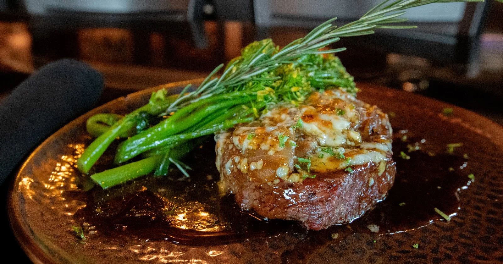 Steak topped with garlic butter, accompanied by green vegetables garnished with rosemary, on a metallic plate.