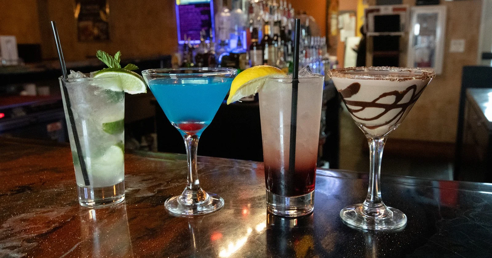 Four colorful cocktails on a bar counter, including a mojito, a blue martini, a layered pink and red drink, and a chocolate drizzle martini.