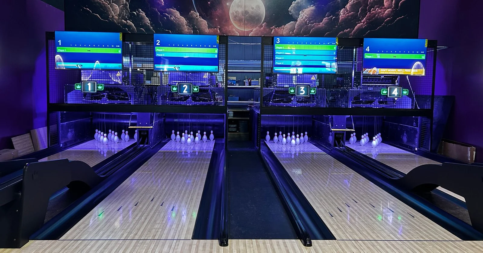 Four bowling lanes with pins set up at the end of each lane. Digital screens displaying scores hang above each lane. The background features a mural of the moon and clouds.