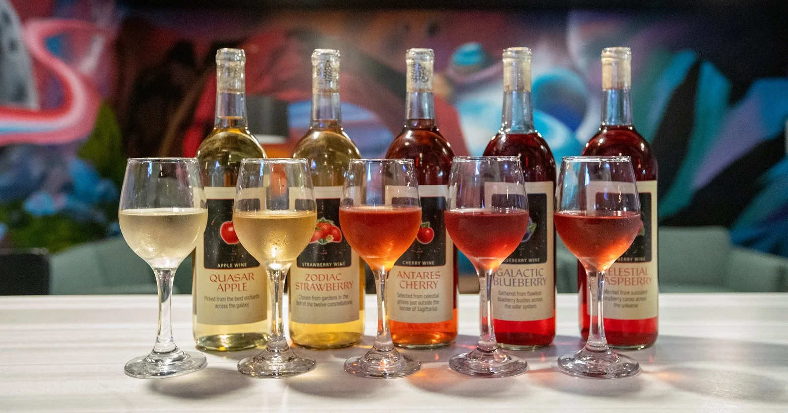 Five glasses of different colored wines in front of five bottles labeled with names of celestial bodies and constellations, arranged on a white surface with a abstract colorful mural in the background.