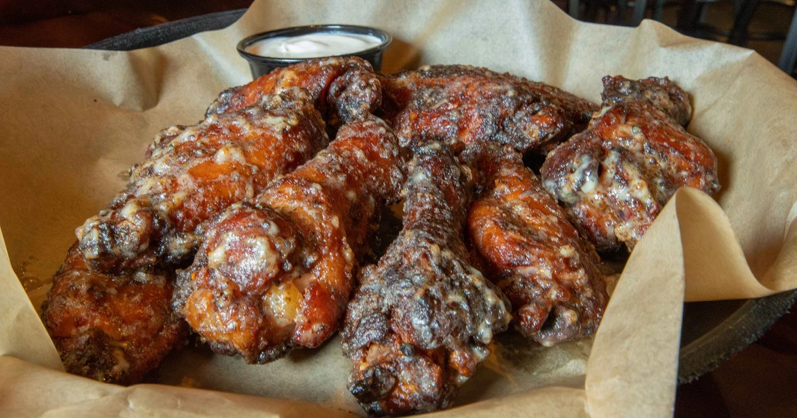 Basket of cooked chicken wings with a side of dipping sauce in a black cup, served on parchment paper.