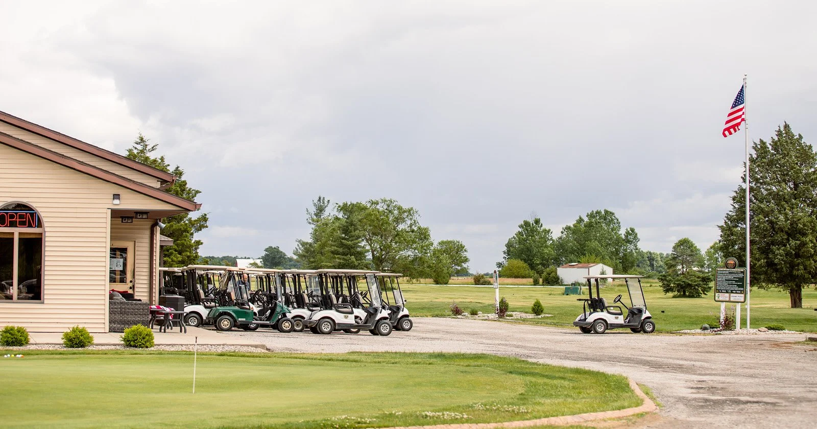 Golf course clubhouse with a line of golf carts parked outside, a putting green in the foreground, and an American flag flying near the building on a cloudy day.