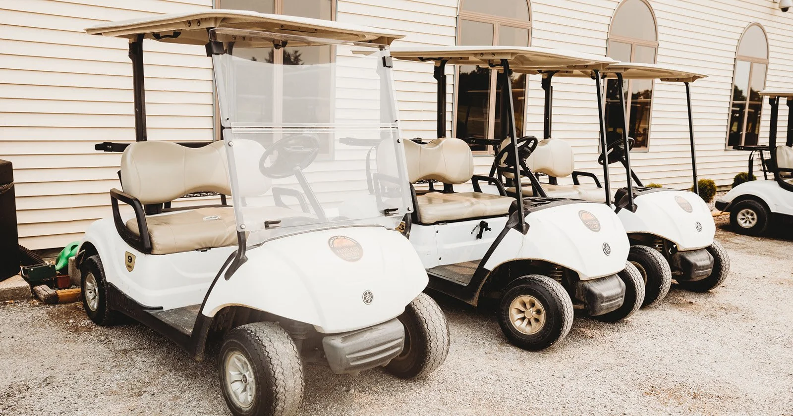 Four white golf carts with beige seats parked outside a building with white siding and arched windows.