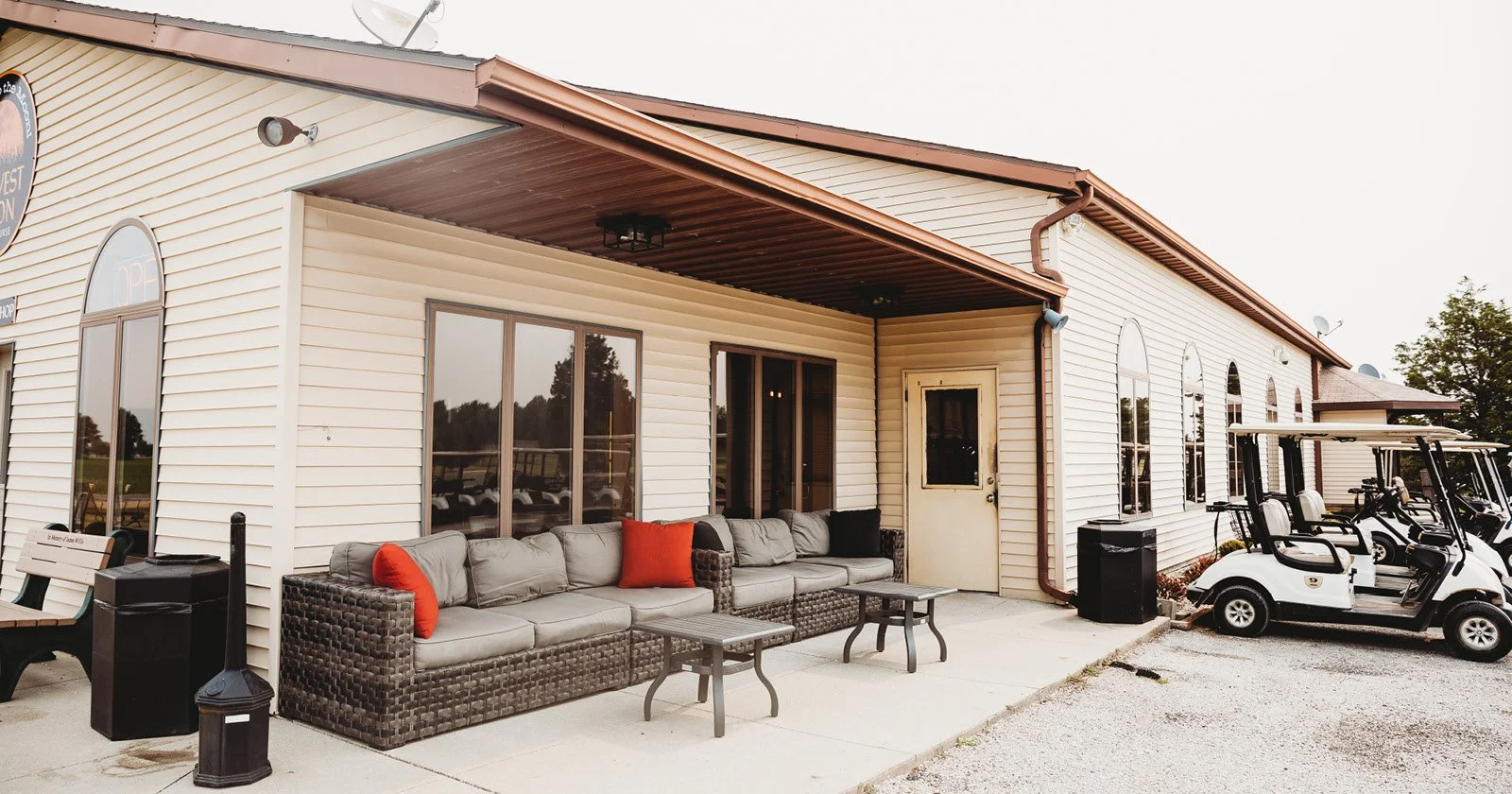 Outdoor patio area with a wicker couch with cushions and red pillows, several small tables, a black trash bin, golf carts parked to the right, and a building with large arched windows and beige siding.