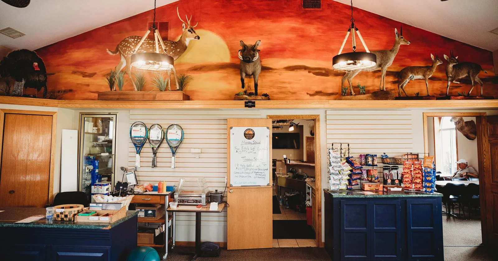 Interior of a restaurant or cafe with a large mural of wildlife, including a deer, antelope, and a mountain goat, on the upper wall painted in sunset colors. The space features a counter area with tennis rackets hanging on the wall, snack stacks, and