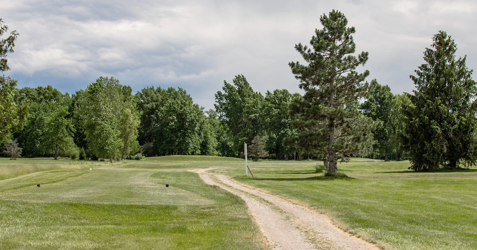 A dirt path runs through a lush, green golf course bordered by trees, under a partly cloudy sky.