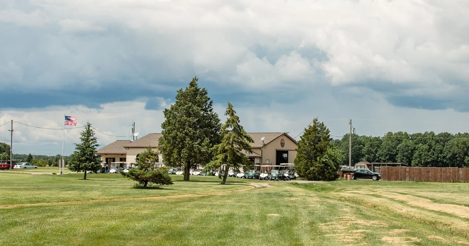 A large open grassy area with a few trees in front of a brown building that appears to be a clubhouse or similar structure, with cars parked nearby. An American flag is flying on a pole on the left side of the image. The sky is partly cloudy with dar