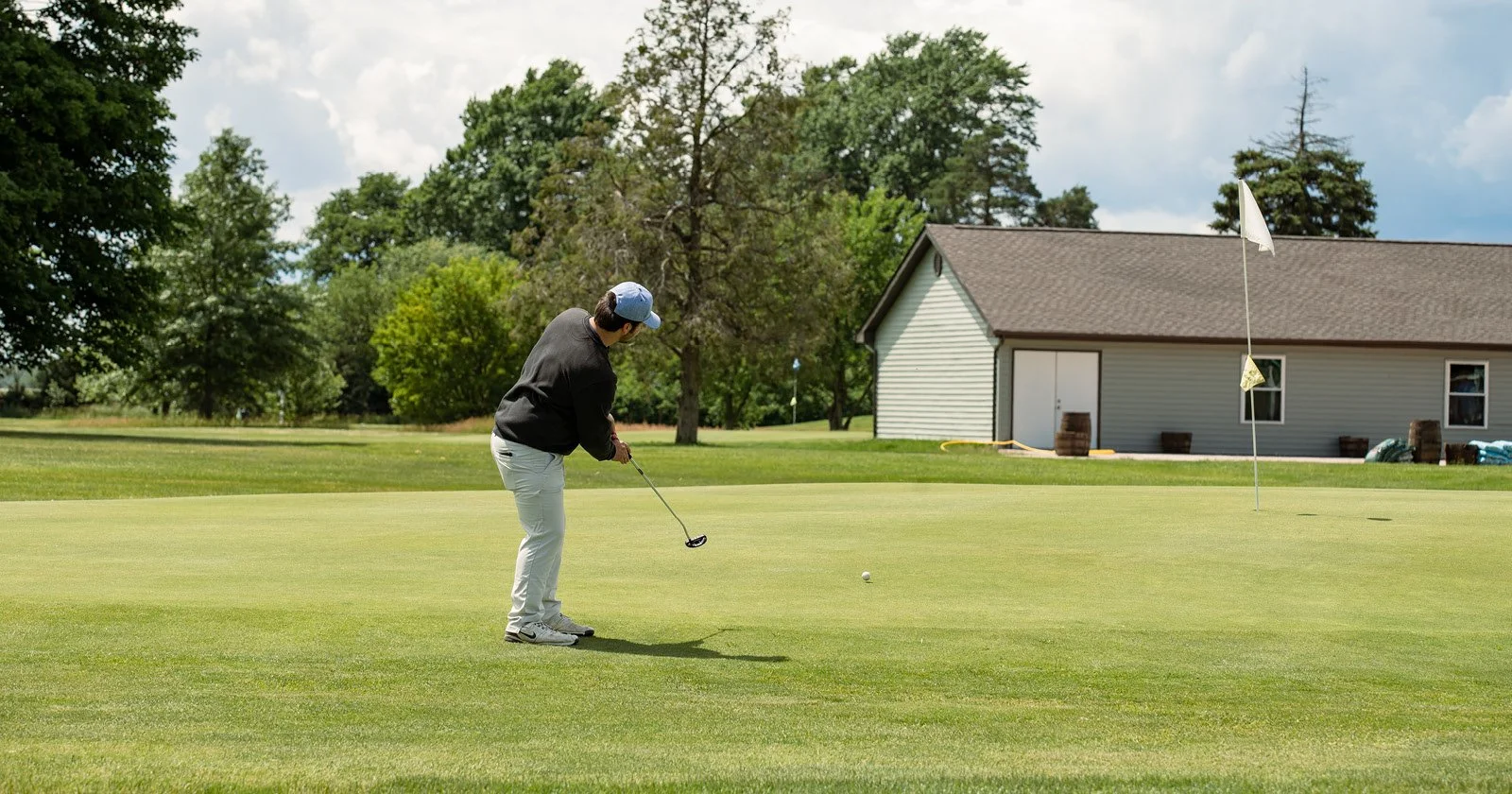 A person in a black shirt and white pants playing golf on a green field near a house, with trees and a cloudy sky in the background.