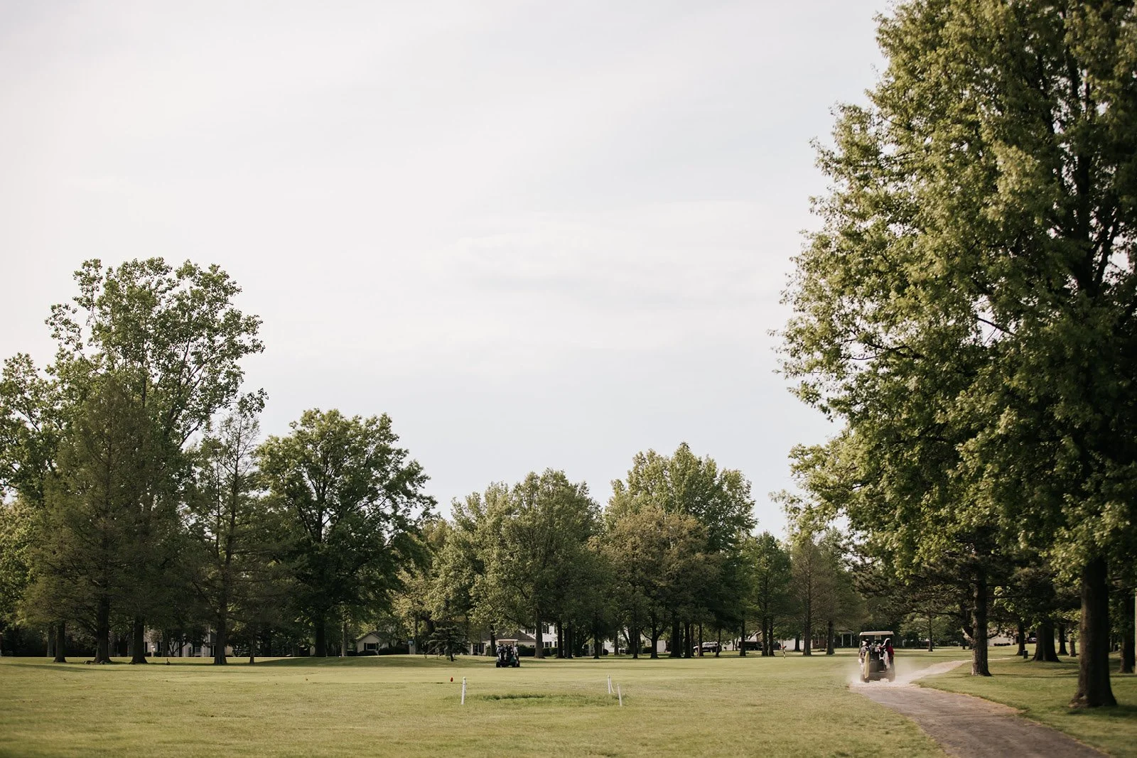 Golf course with trees lining a dirt cart path, several golf carts in the distance, and a partly cloudy sky.