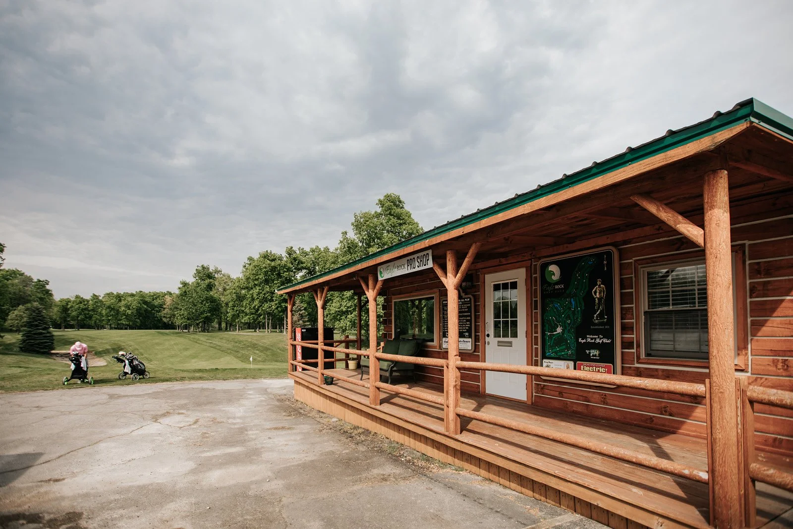 Exterior of a wooden pro shop building at a golf course with a porch, two golf bags on carts, and a grassy area with trees in the background under a cloudy sky.