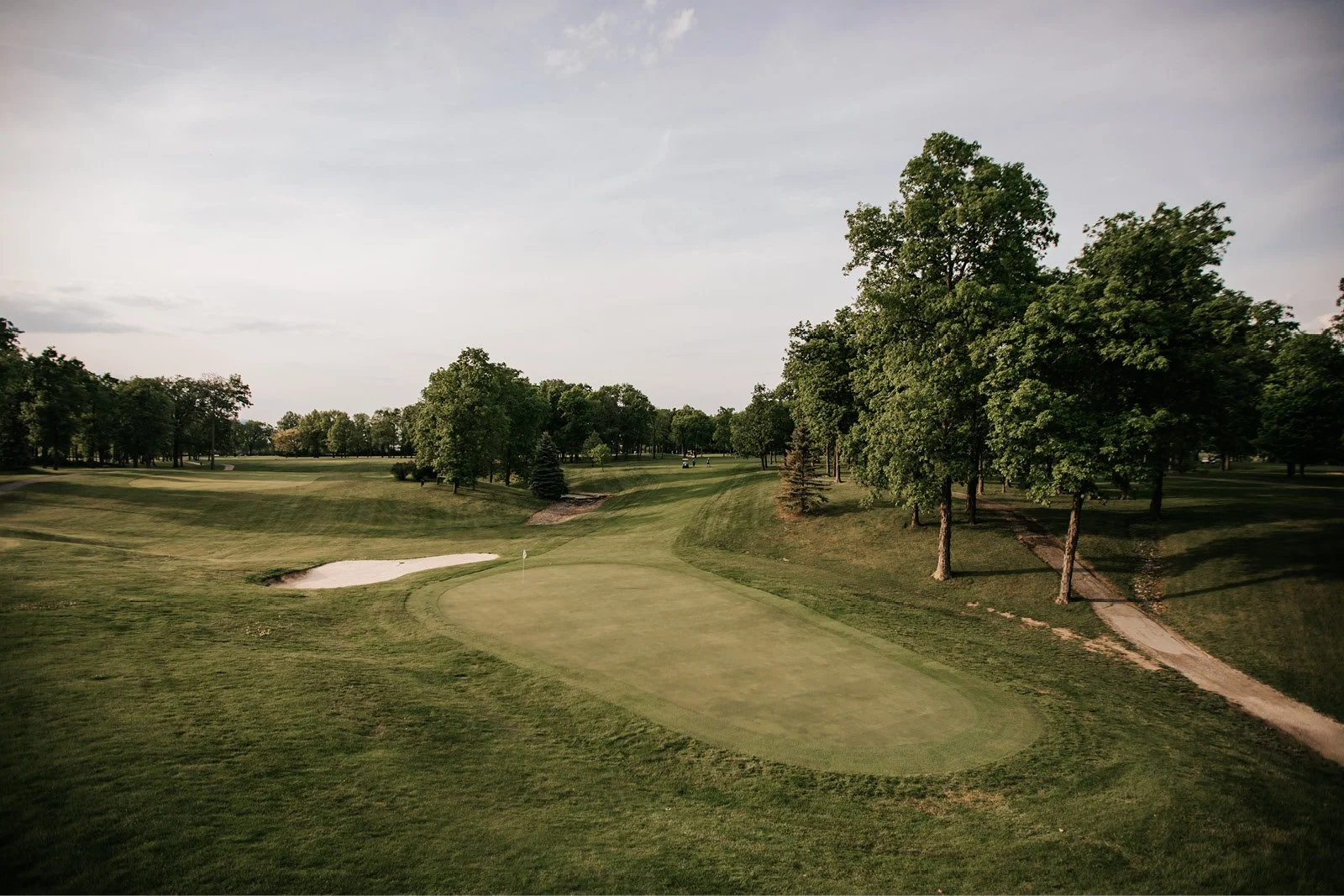 Golf course with green, sand bunker, and trees under a cloudy sky.