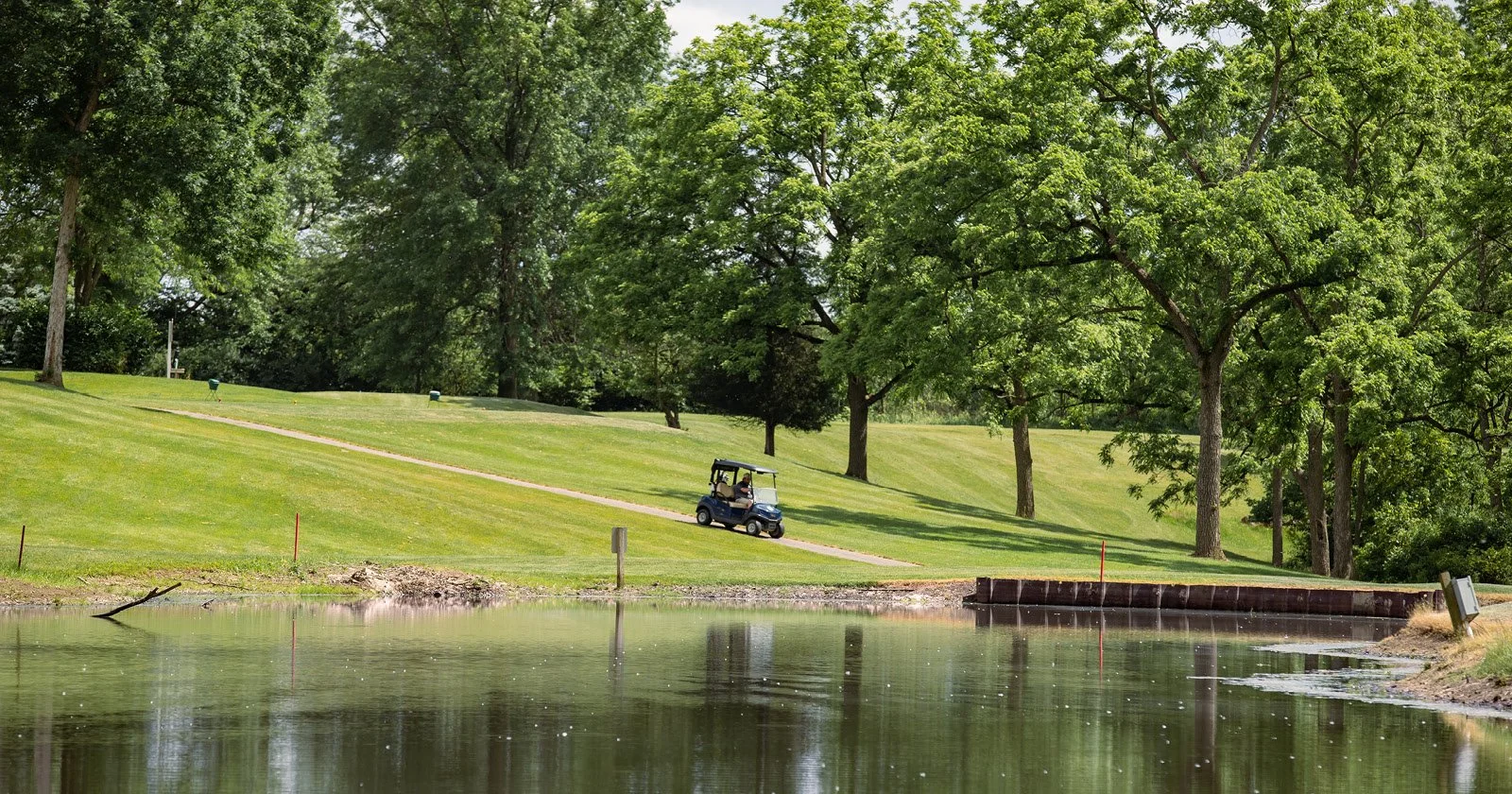 A golf course with a water hazard in the foreground, a golf cart driving on a paved path, and lush green trees in the background.