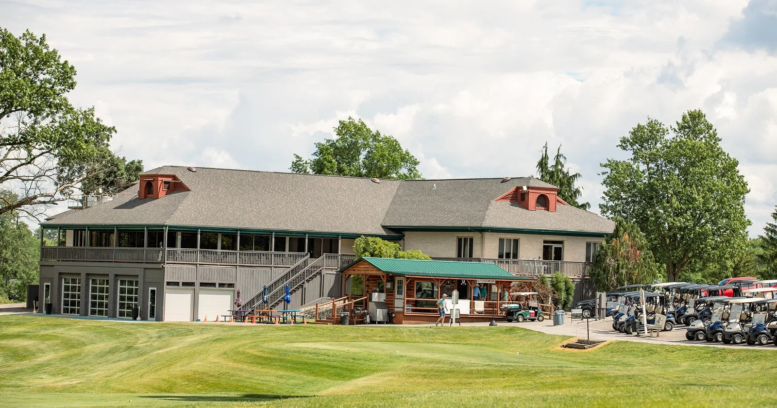 A large building with a gray roof and white exterior walls, surrounded by lush green trees and a well-maintained grassy area. There are several golf carts parked on the right side and a small wooden structure with a green roof in front of the buildin