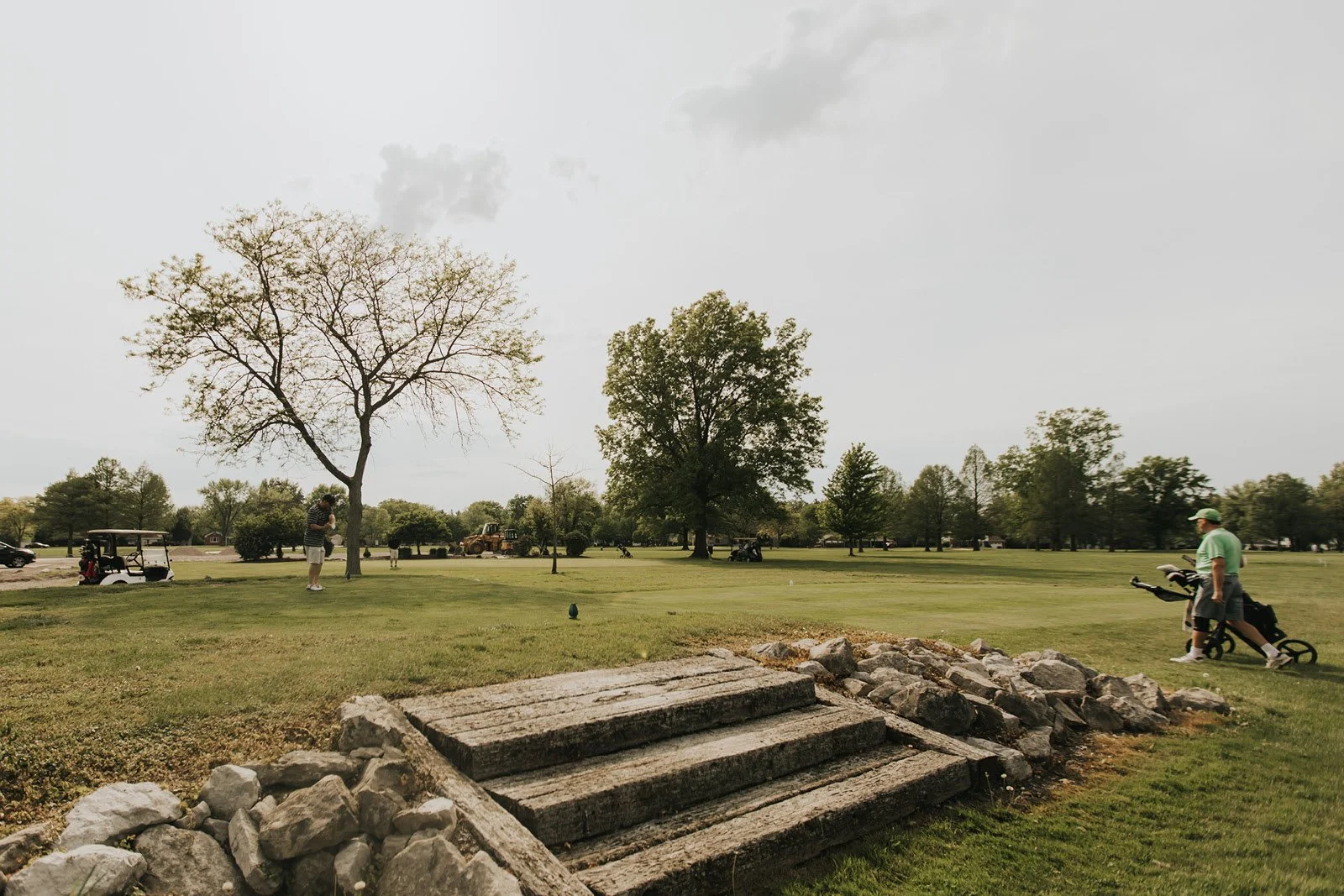 Golf course with people playing, trees, and cloudy sky.