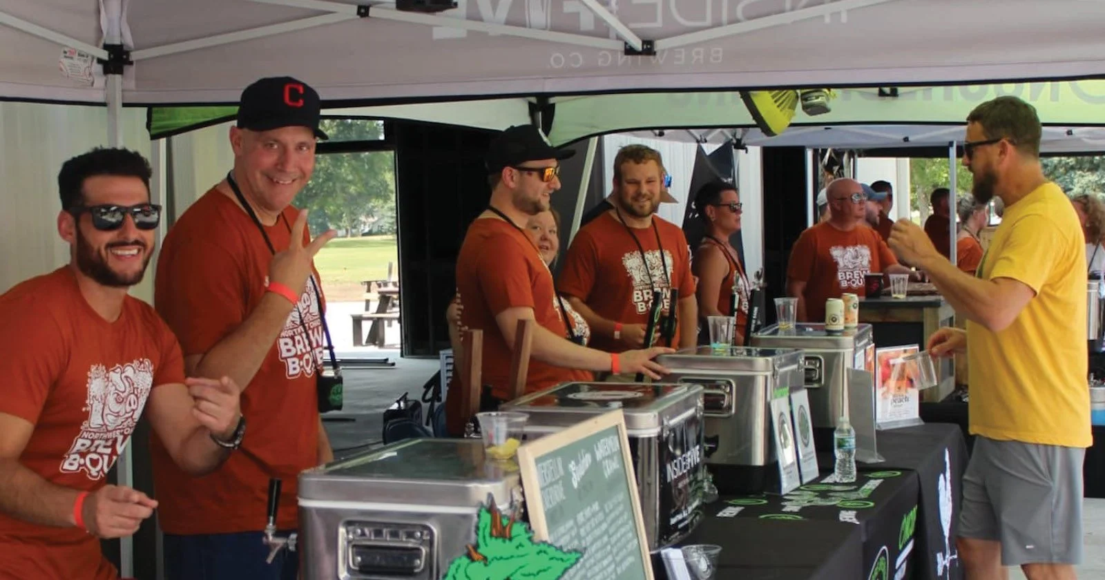 Group of smiling men at a beer tent, some making peace signs, with a man in a yellow shirt ordering drinks from the staff. The staff members are wearing matching orange shirts and are standing behind a counter with coolers and promotional flyers.