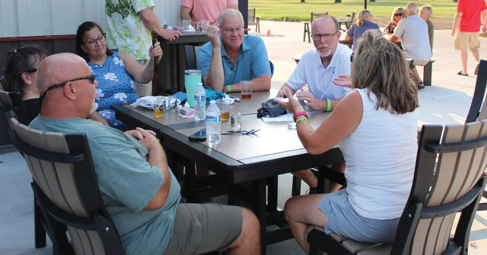 A group of six adults sitting around a rectangular outdoor table, engaged in conversation during a social gathering. Some are wearing glasses, and there are drinks and personal items on the table. In the background, other groups are seated at tables