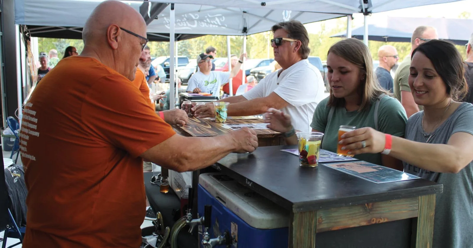People at an outdoor beer tent, with a man pouring beer from a tap into a glass, patrons holding drinks and menus, and parked cars in the background.