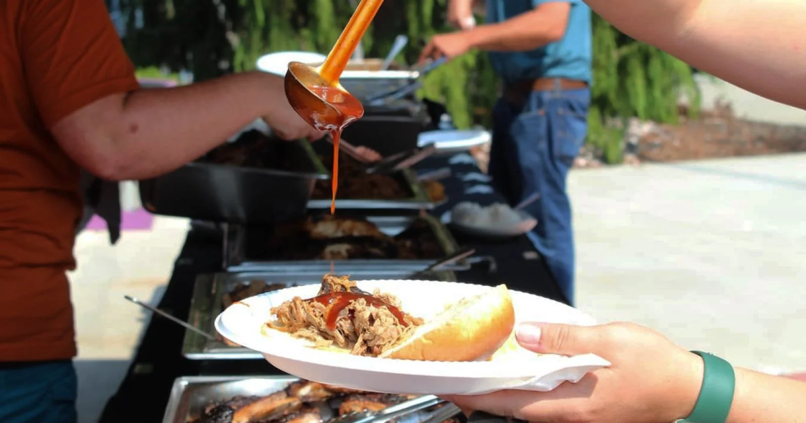 Person holding a paper plate with a pulled pork sandwich, topped with barbecue sauce from a ladle at an outdoor food event with a buffet of dishes in the background.