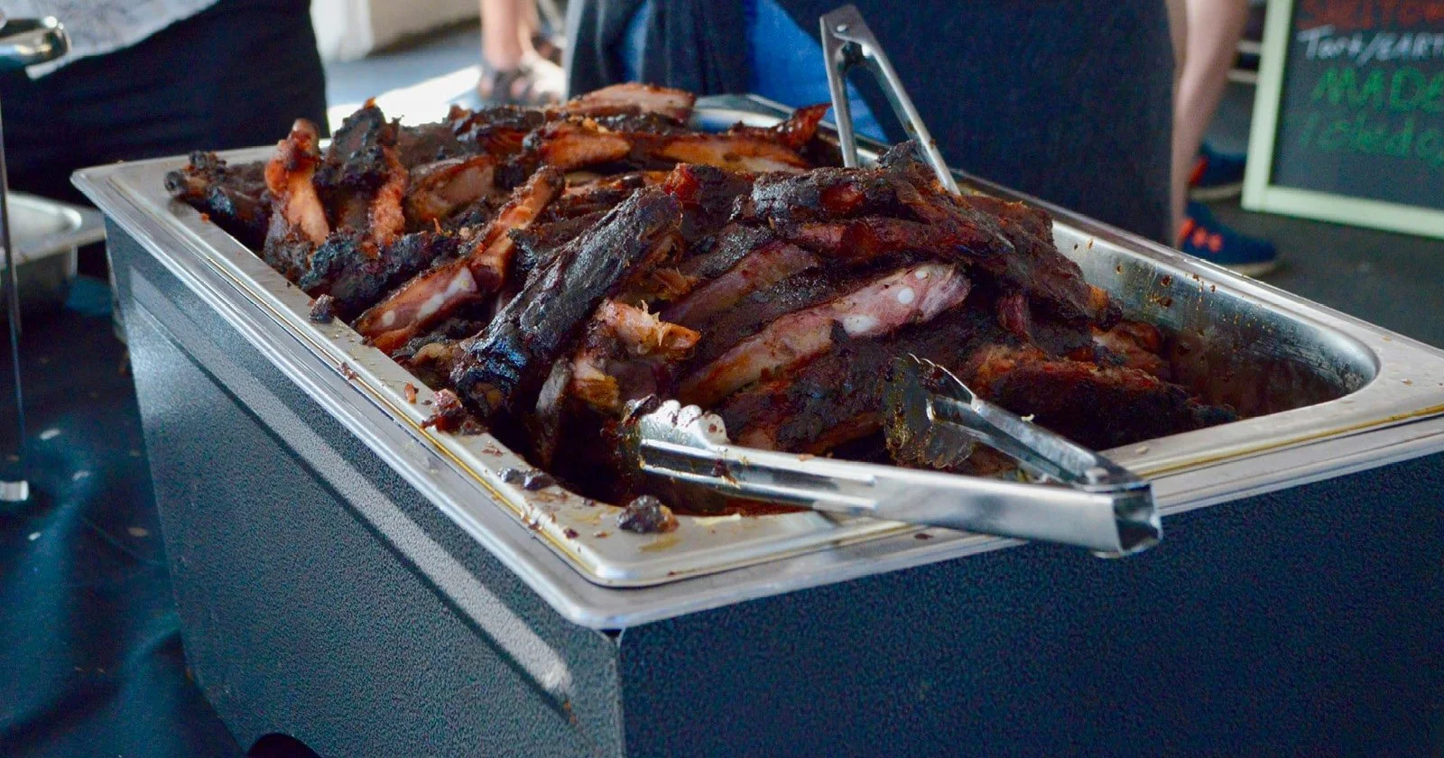A tray of cooked, smoky barbecue ribs with tongs on a table at a food event.