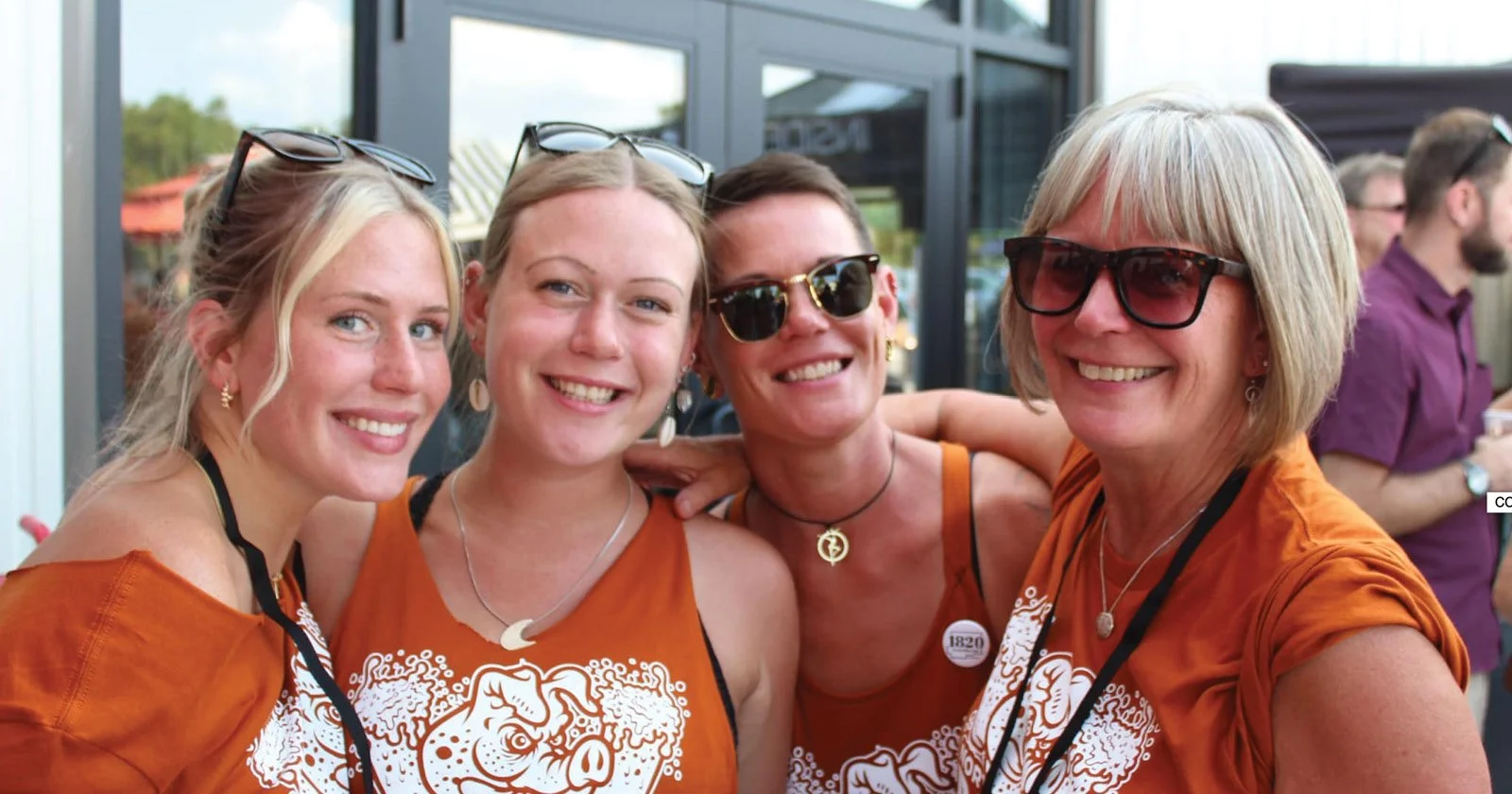 Four women smiling and wearing matching orange shirts, standing close together outside near a glass building, with other people in the background.