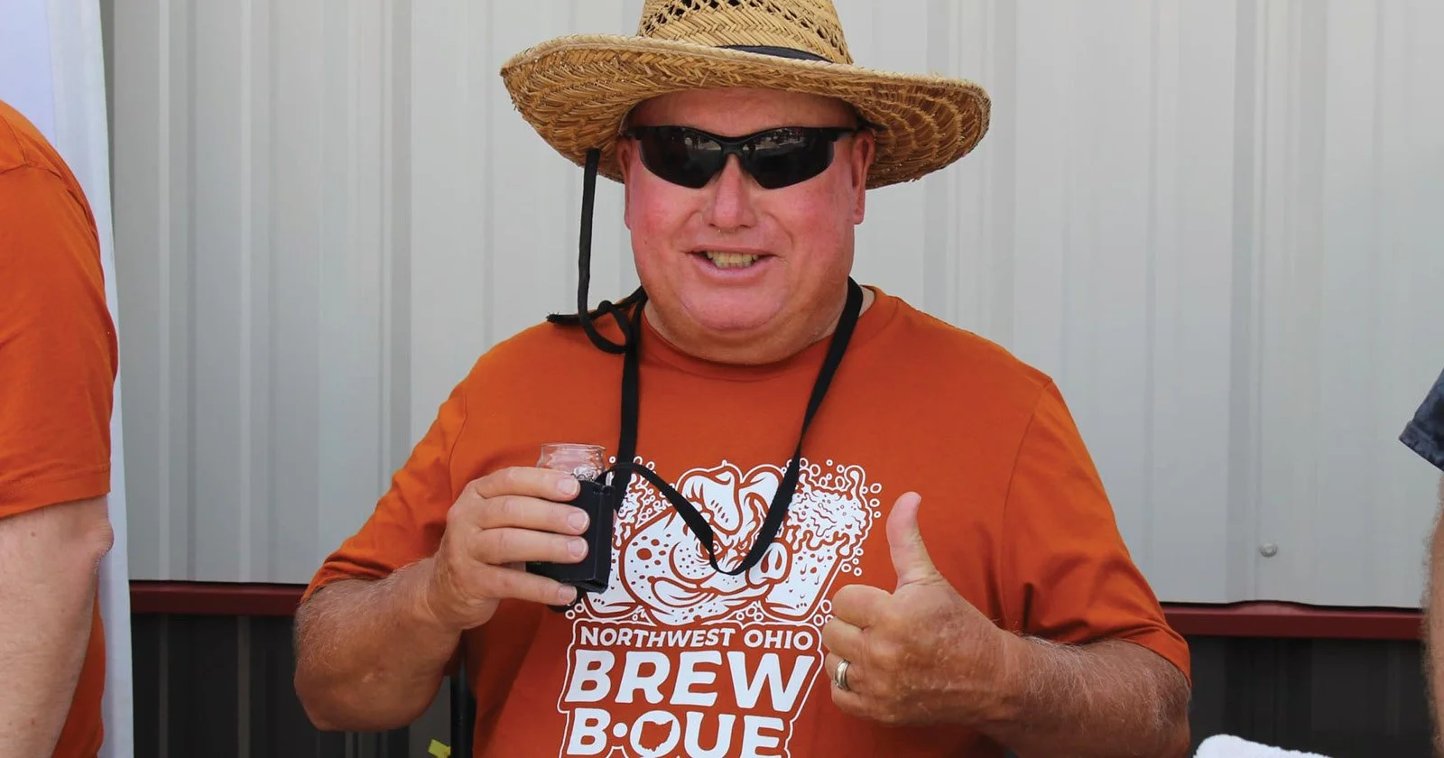 A man wearing a straw hat, sunglasses, and an orange T-shirt with a logo and text, holding a small glass of dark beverage, giving a thumbs-up gesture, in front of a light-colored wall.