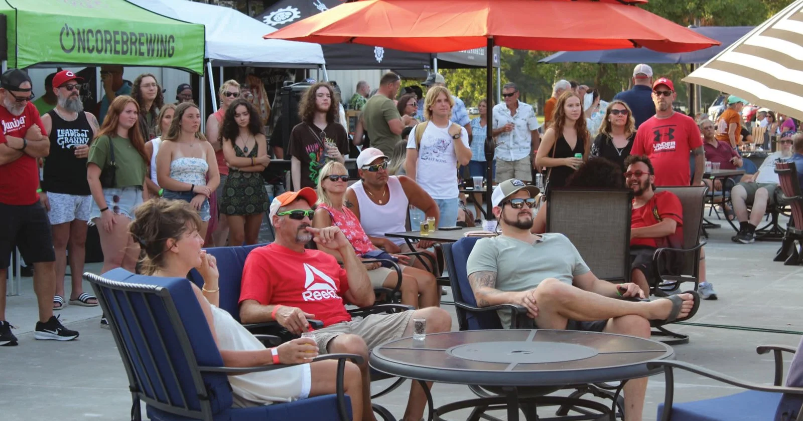 People gathered at an outdoor event or festival with seating, umbrellas, and vendor tents, including a green tent labeled 'ONCEREBRWING' and a striped umbrella visible in the background.