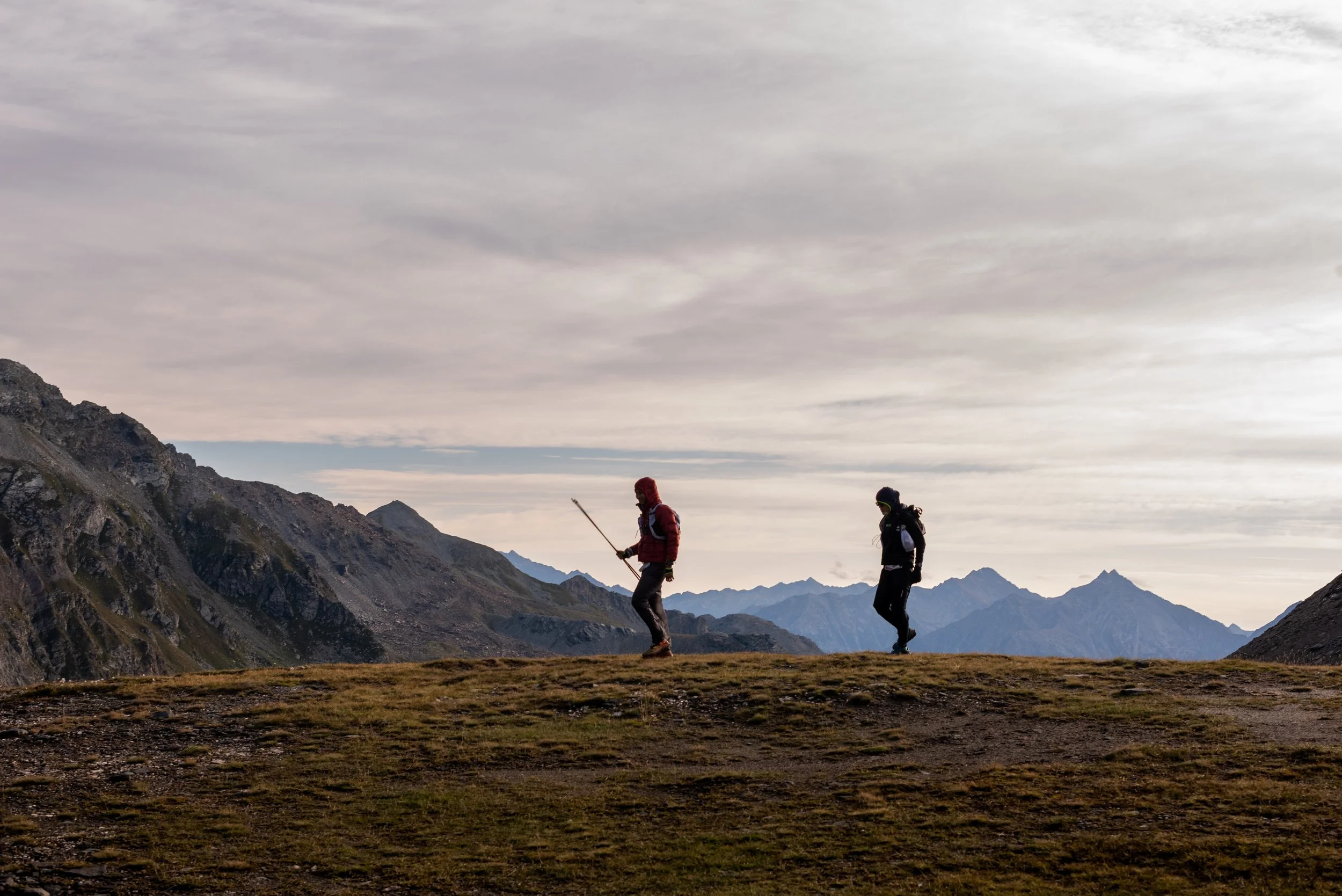Due escursionisti in montagna durante il tramonto, uno con bastone da trekking, sullo sfondo catene montuose e cielo nuvoloso.