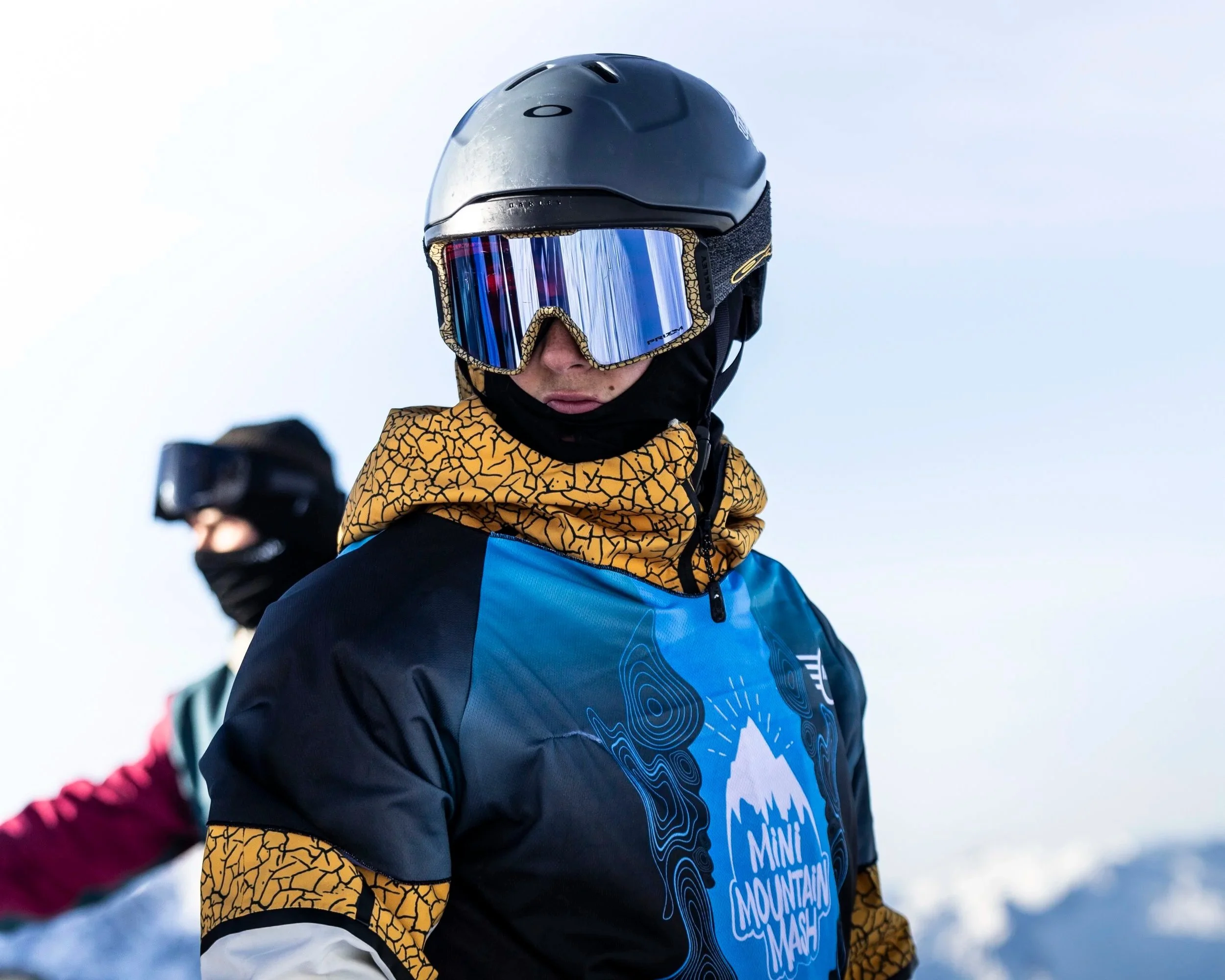 Due persone sulla neve con attrezzatura da sci, una in primo piano indossa casco, maschera da sci, giacca blu con dettagli gialli, e un maglione con il logo 'Mini Mountain Mash'. L'altra persona è sullo sfondo, anch'essa con casco e maschera, e giacc