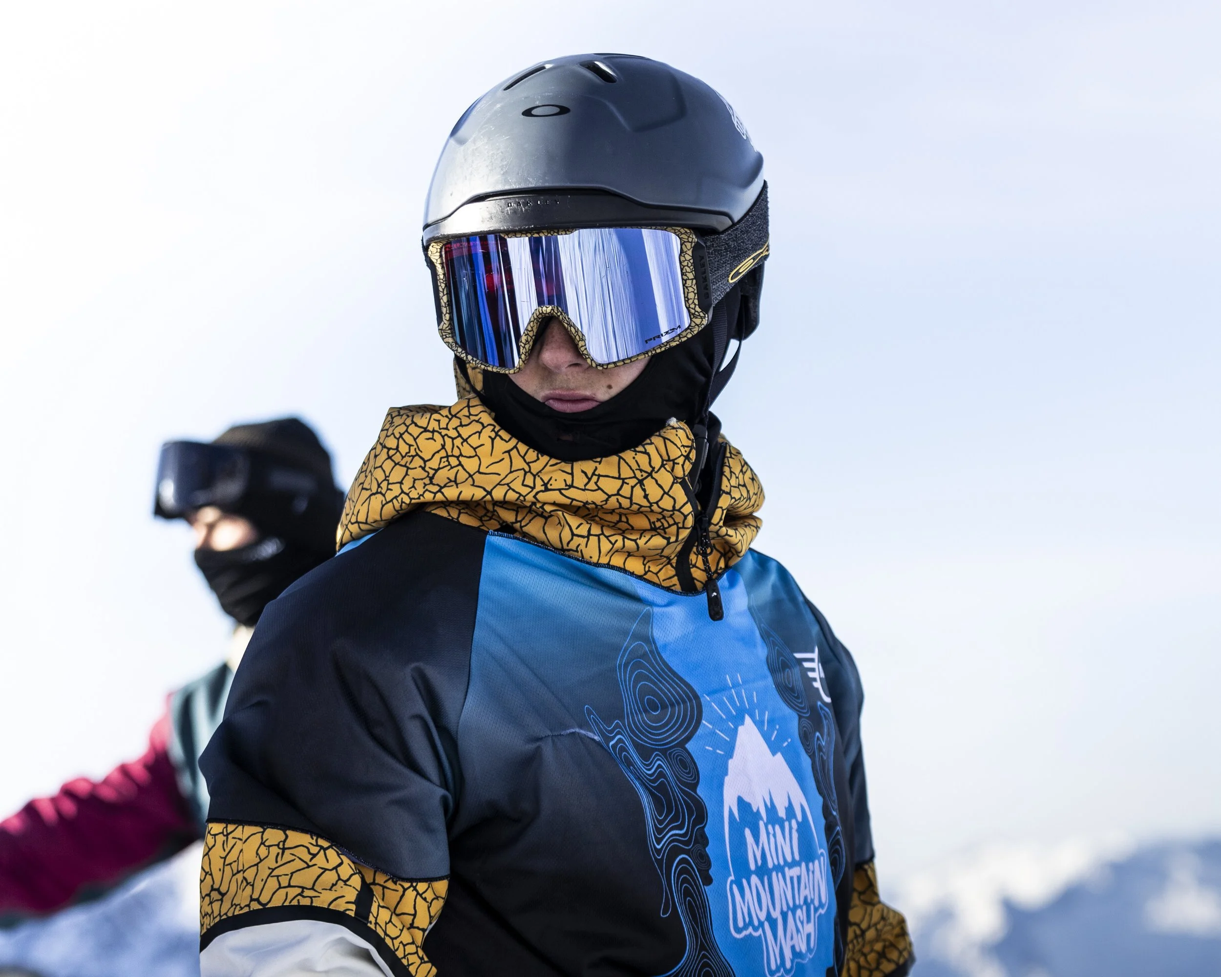 Due persone sulla neve con attrezzatura da sci, una in primo piano indossa casco, maschera da sci, giacca blu con dettagli gialli, e un maglione con il logo 'Mini Mountain Mash'. L'altra persona è sullo sfondo, anch'essa con casco e maschera, e giacc