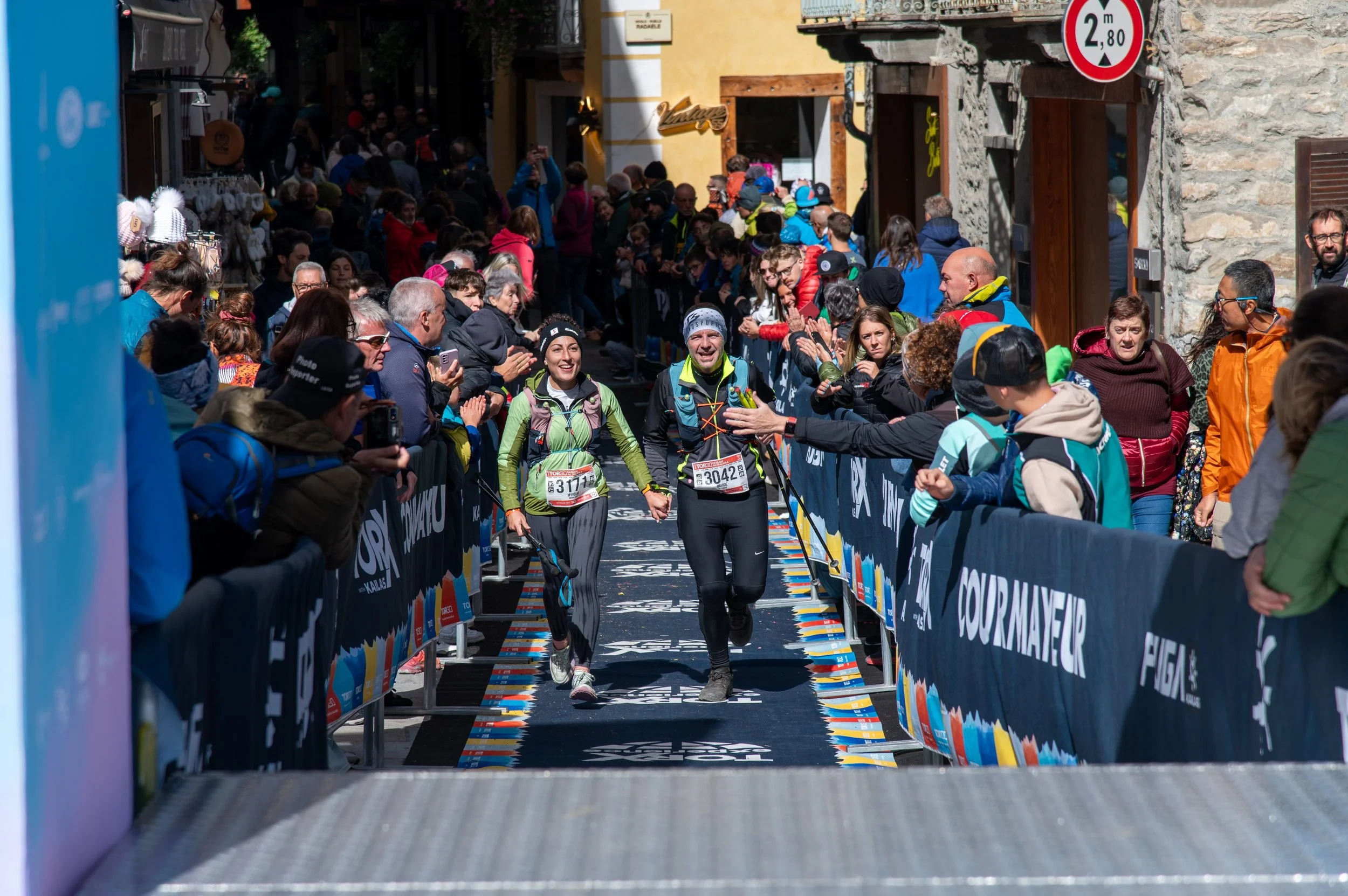 Due atleti sorridenti che attraversano il traguardo di una maratona, circondati da una folla di spettatori entusiasti, su una strada cittadina con bar e edifici antichi.
