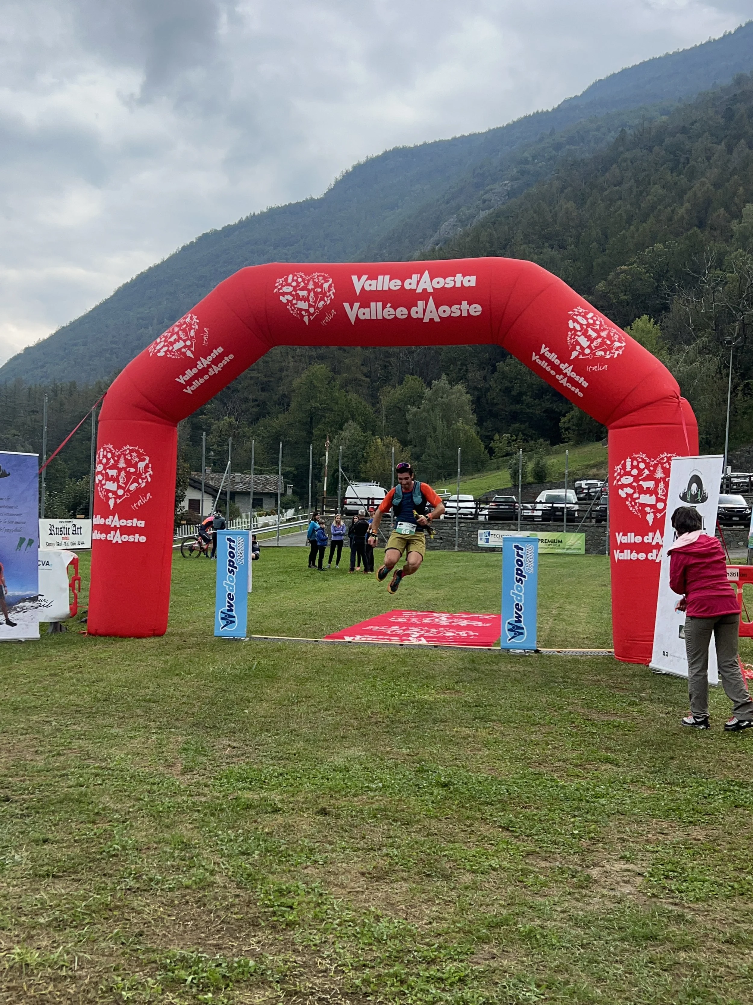 Un uomo sta partecipando a una corsa o gara di arrampicata, sta saltando sopra una linea di partenza o arrivo sotto un arco rosso con scritte 'Valle d'Aosta' e 'Vallée d'Aoste', in un'area all'aperto con montagne sullo sfondo.