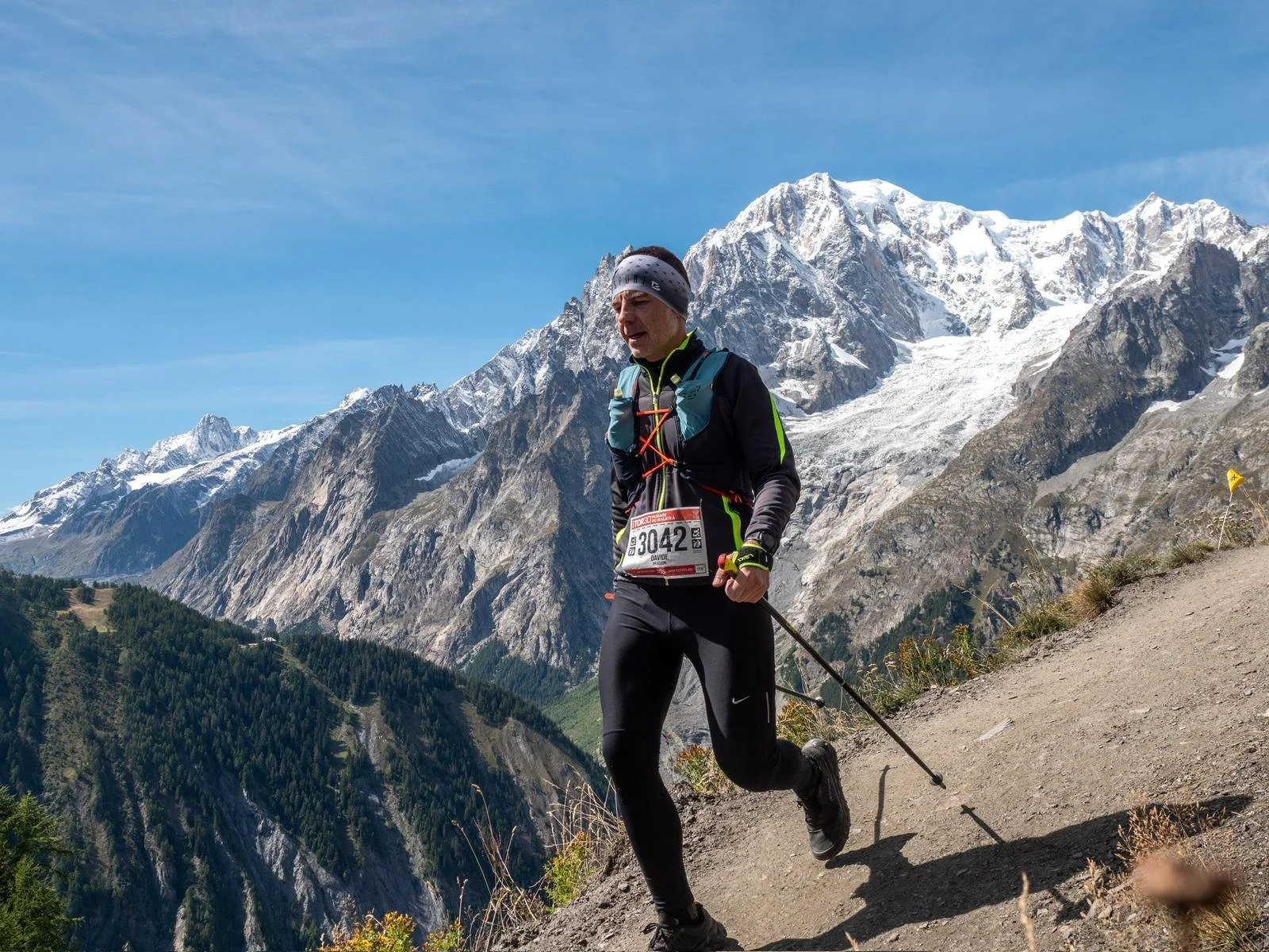 Uomo che corre in montagna con scarpe da trekking e bastoncini, sfondo di montagne con neve e cielo blu.