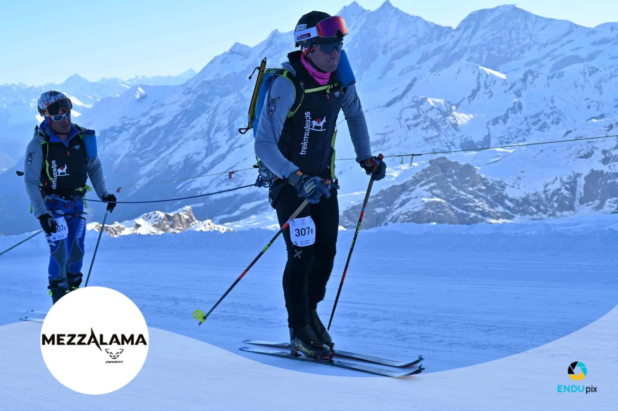Due scaler in allenamento su una superficie innevata con montagne innevate sullo sfondo, indossando abbigliamento sportivo e attrezzatura da sci.