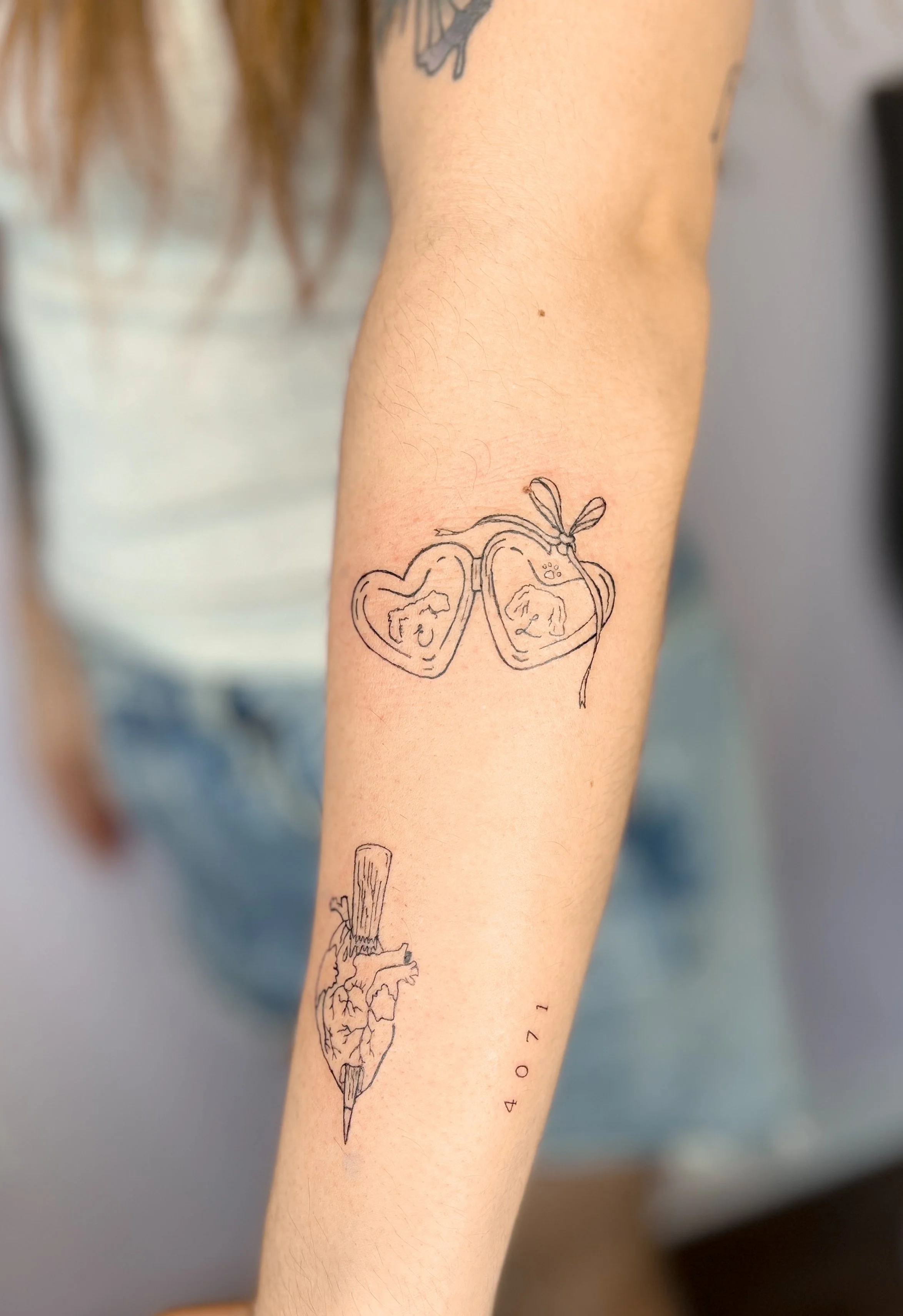 Line art tattoo of heart with a wooden stake, a locket with two dogs, and a small date written vertically below.
