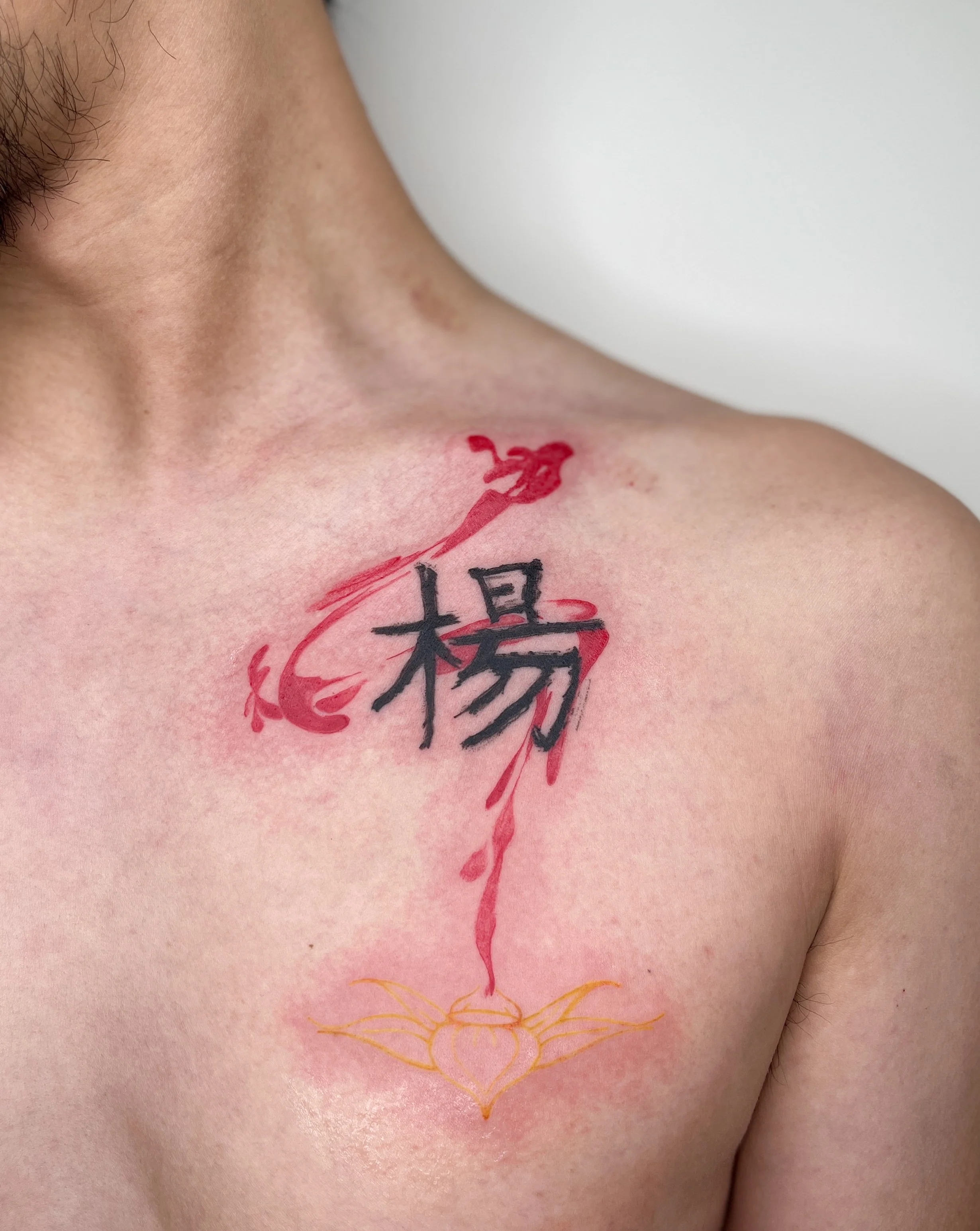 Tattoo of a black Chinese character with red ink and orange decorative design on a person's chest and shoulder.