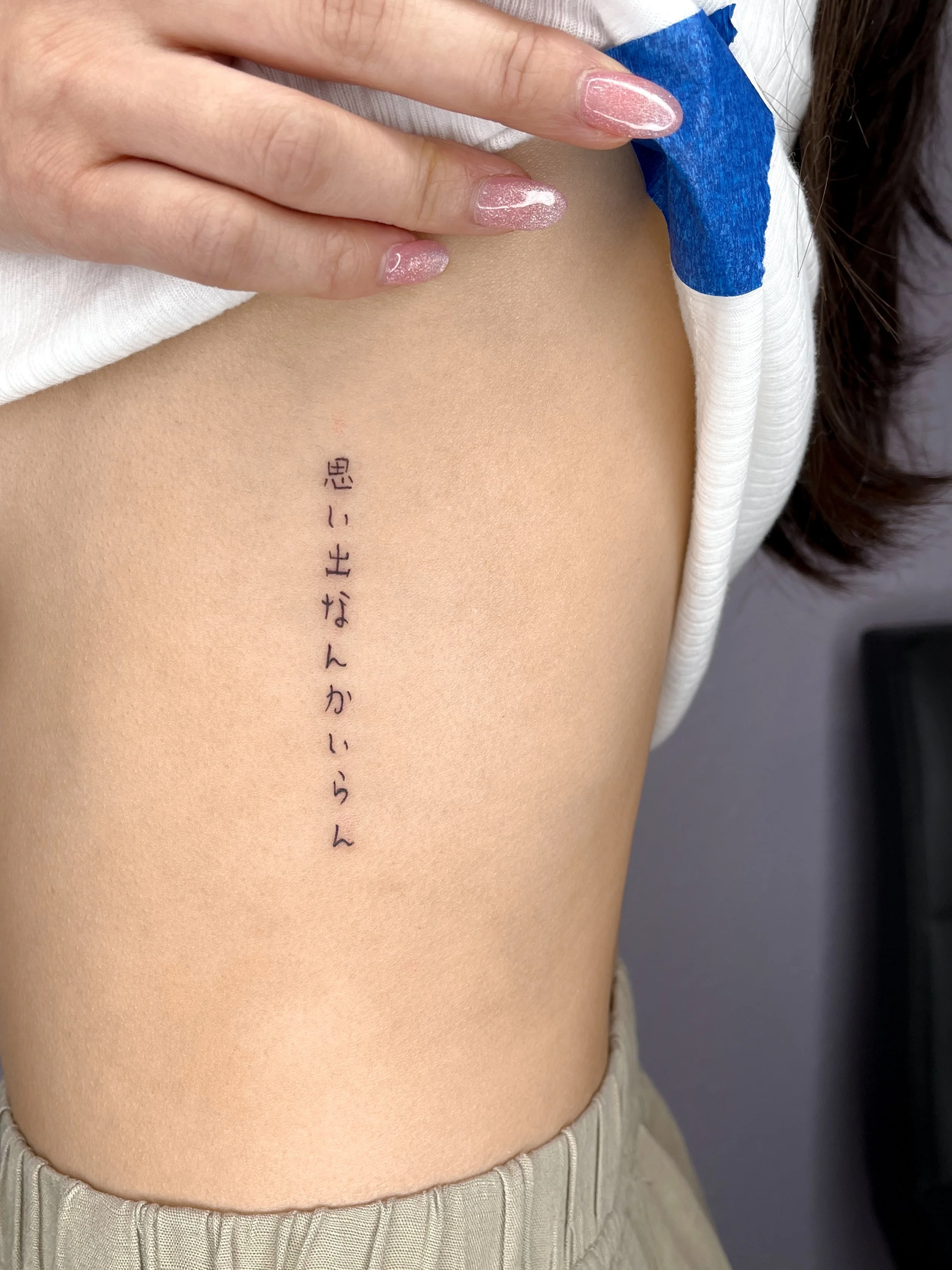 A person's side showing a fresh, black ink tattoo of Japanese characters on their ribcage, partially covered by a white shirt with blue tape.