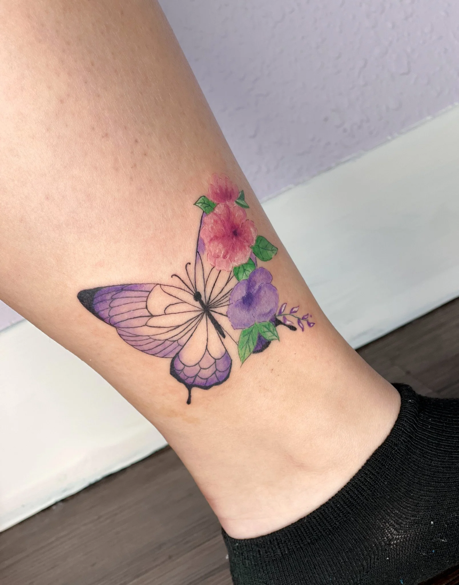 Tattoo of a purple and white butterfly with pink and purple flowers and green leaves on a person's leg.