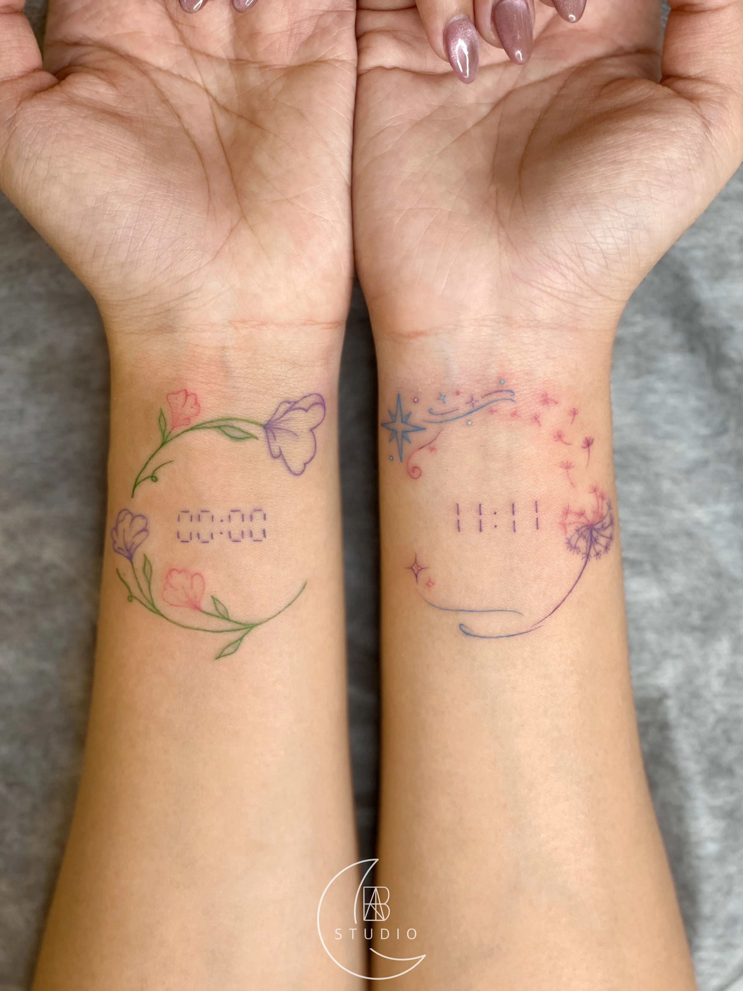 fine linework tattoo in pastel colors; 00:00 and 11:!1 reflecting each other of the wrists with one side in a floral design and the other in a starry night design 